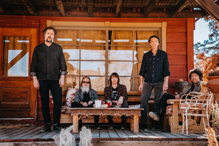 The bands Drive-By Truckers and Deer Tick pose on a rustic wooden porch with relaxed expressions.