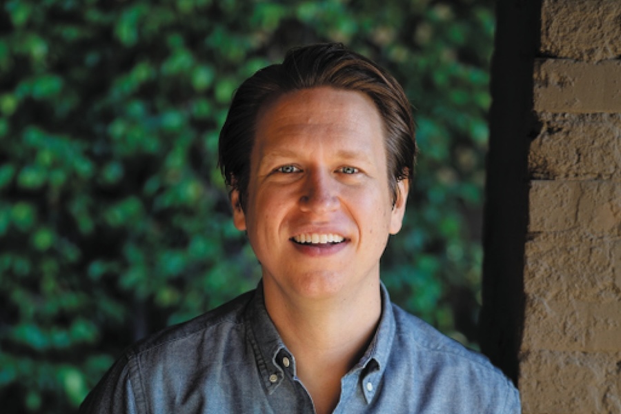 Comedian Pete Holmes smiles while standing against a leafy background and brick wall.