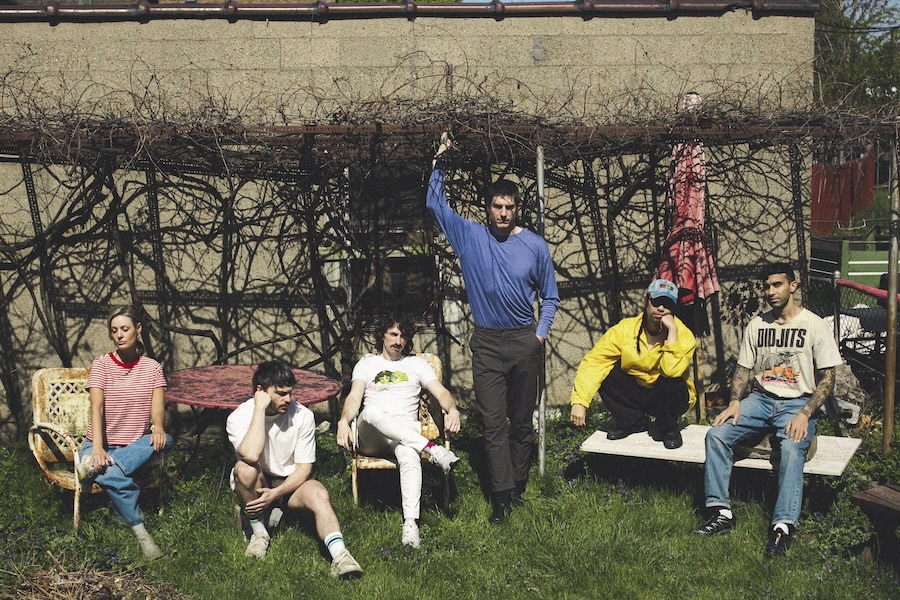 The Armed band members pose in an overgrown backyard with vintage chairs and a vine-covered trellis.