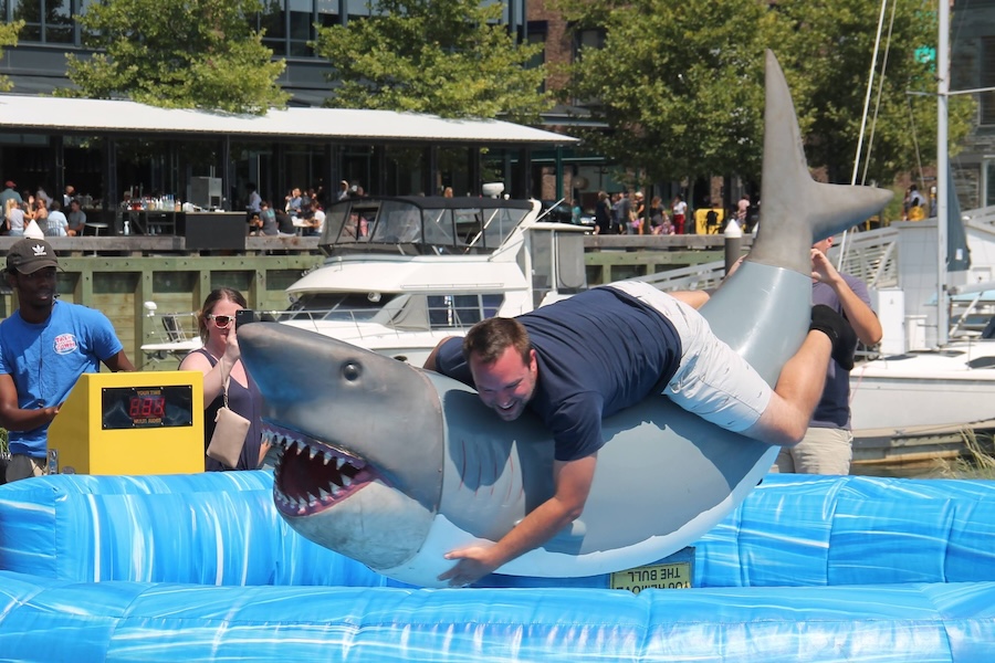 A man clings to a mechanical shark ride during an outdoor waterfront event.